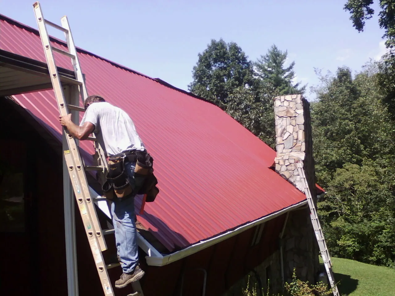 Roofer on a ladder beside a red metal roof and stone chimney — Soffit & Fascia Repair by Roofing Brothers in Elm City