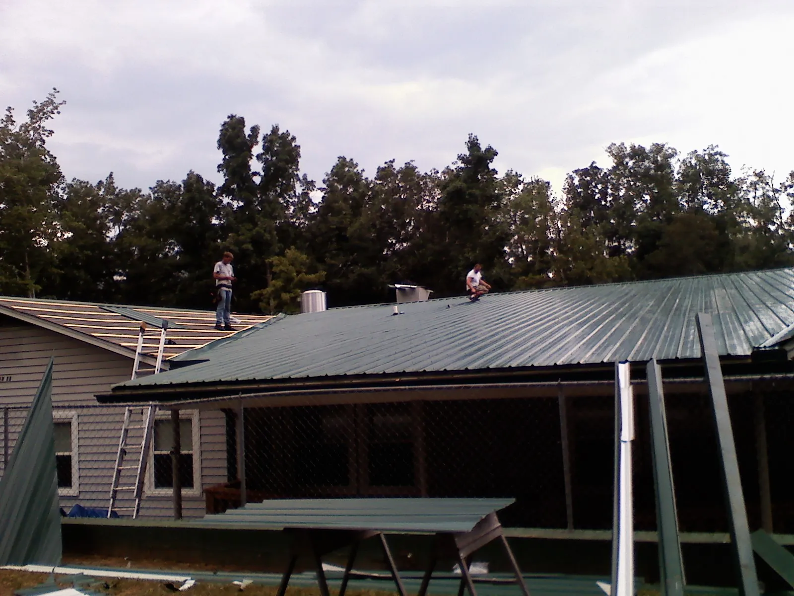 Two roofers installing green metal panels above a covered porch in Elm City